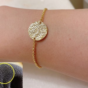 Könnte beinhalten: Ein goldfarbenes Armband mit einem strukturierten, kreisförmigen Anhänger. Der Anhänger ist an einer feinen Kette befestigt. Das Armband wird an einem Handgelenk getragen, wodurch das Design des Schmucks hervorgehoben wird. Die Textur des Anhängers ähnelt einer Nahaufnahme der Hundenase.