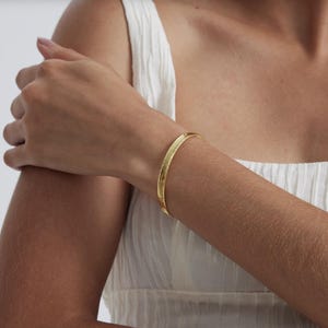 Op de afbeelding: Een goudkleurige armband met een getextureerd oppervlak. De armband wordt om een arm gedragen en de achtergrond is een witte top.