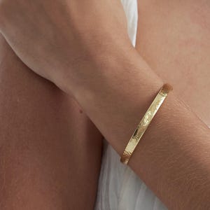 Op de afbeelding: Een goudkleurige armband met gegraveerde tekst. De armband heeft een gehamerde textuur en wordt om een arm gedragen. Een eenvoudig, elegant sieraad, geschikt voor dagelijks gebruik.