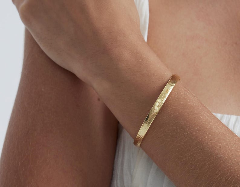 Op de afbeelding: Een goudkleurige armband met gegraveerde tekst. De armband heeft een gehamerde textuur en wordt om een arm gedragen. Een eenvoudig, elegant sieraad, geschikt voor dagelijks gebruik.