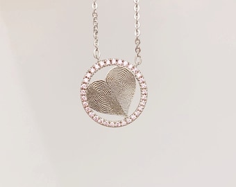 Puede incluir: Un collar de plata con un colgante circular que presenta un diseño de huella digital en forma de corazón rodeado por un anillo de pequeñas piedras preciosas rosas.
