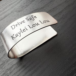 May include: A silver-colored metal cuff bracelet with the engraved text "Drive Safe Kaylei Lou Lou". The bracelet has a curved design and is resting on a dark, textured surface.