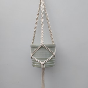 Pode incluir: Um suporte de plantas de macramé branco com um vaso de cerâmica cinza claro listrado. O suporte é feito com um cordão de algodão grosso e tem uma franja na parte inferior.