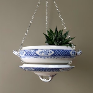 May include: A hanging planter made from a white ceramic bowl and matching saucer with blue floral patterns. The bowl features two side handles and is suspended by three silver-toned metal chains. A green succulent plant sits inside the bowl.