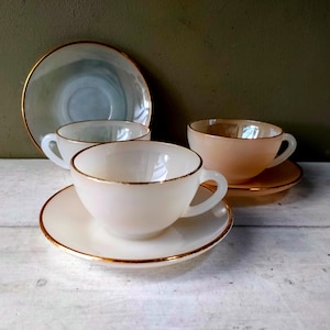 May include: A collection of vintage teacups and saucers. The cups and saucers are in shades of white and peach, with gold rims. One saucer is a light blue-grey color. The set is arranged on a white wooden surface.