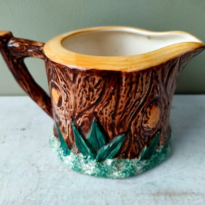 Puede incluir: Una jarra de crema de cerámica marrón con forma de tronco de árbol con hojas verdes alrededor de la base. La jarra tiene un borde marrón claro y un interior blanco.