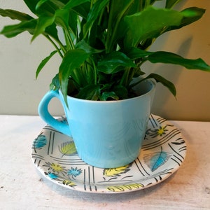 May include: A light blue teacup with a handle, filled with a green plant, sits on a matching saucer. The saucer has a white background with a black, yellow, and blue leaf pattern. The teacup and saucer are a decorative plant pot.