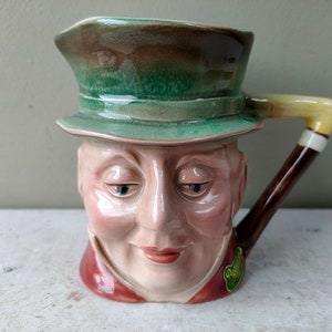 May include: A ceramic pitcher shaped like a man's head wearing a green hat. The pitcher has a brown handle shaped like a pipe.