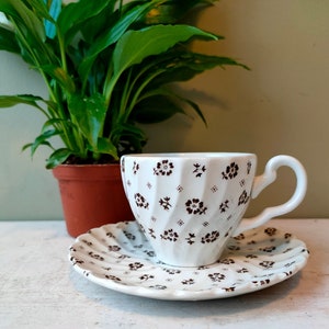 May include: A white teacup and saucer with a brown floral pattern. The cup has a fluted design and a handle. The saucer has a scalloped edge.
