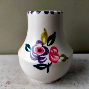 Puede incluir: Un pequeño jarrón de cerámica blanca con cuerpo redondeado y parte superior acampanada. Decorado con flores pintadas a mano en tonos morado, rosa, amarillo y verde. El borde está adornado con pequeños puntos azul oscuro.