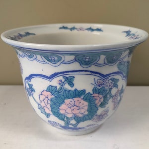 May include: A white ceramic planter with a blue and green floral design. The planter has a wide rim and a rounded base.