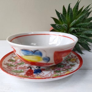 May include: A small, white ceramic bowl and saucer set with a hand-painted design. The bowl has red, blue, and yellow accents. The saucer features a detailed floral and figure pattern with a red border. A succulent plant is in the background.