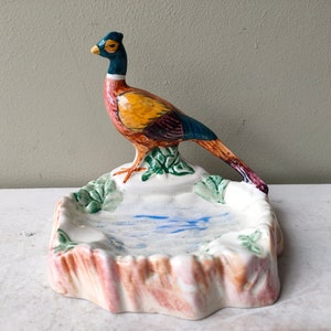 May include: A ceramic trinket dish with a colorful pheasant figurine perched on the edge. The dish is decorated with a blue and white painted pond and green foliage.
