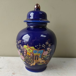 May include: A cobalt blue ceramic jar with a lid, featuring a decorative scene of a carriage and floral elements. The jar has a glossy finish and a gold-colored finial on the lid. The jar is a decorative item.
