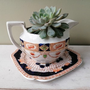 May include: A white porcelain pitcher with a blue and gold floral pattern, used as a planter for a succulent plant. The pitcher is sitting on a matching saucer.