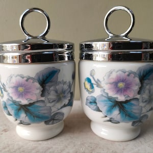 Puede incluir: Dos coquetieras de cerámica blanca con tapas plateadas y diseños florales. El diseño floral presenta flores azules, moradas y rosas con hojas verdes.