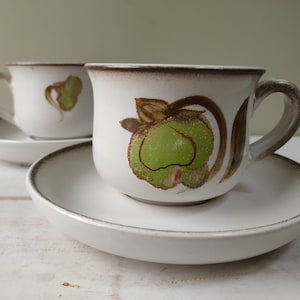 Puede incluir: Dos tazas de té de cerámica blanca con bordes marrones y diseños florales. Las tazas tienen patrones florales verdes y marrones. Las tazas están sobre platillos blancos.