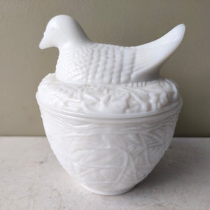 May include: White milk glass bird and nest dish. The bird is perched on a nest with detailed leaves and flowers. The dish is round with a white, textured design.
