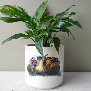 May include: A white ceramic planter with a fruit design featuring a pear, an orange, and a pumpkin. A green peace lily plant is growing in the planter.
