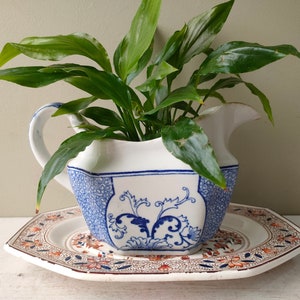 May include: A blue and white porcelain jug with a floral design, used as a planter for a green plant. The jug is sitting on a matching plate with a red and orange floral design.