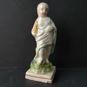 Puede incluir: Una figurita de cerámica blanca de una persona joven de pie sobre una base verde. La figurita lleva una túnica blanca con detalles azules y rojos. La figurita sostiene un objeto amarillo y marrón en su mano derecha.