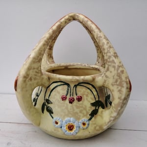 May include: A ceramic planter with a handle, decorated with a floral design in shades of yellow, green, red, and blue. The planter has a speckled glaze and a unique shape.