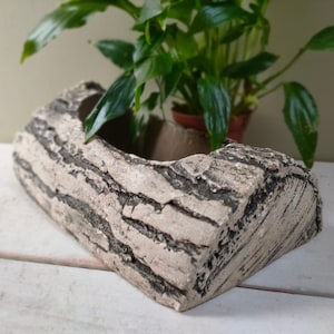 May include: A rectangular planter with a textured, bark-like appearance. The planter is light beige with dark grey streaks, resembling a section of a tree trunk. Green plants are visible in the background, suggesting the planter is used for indoor gardening.