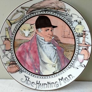 Op de afbeelding: Een wit keramiek bord met een bruine rand en een kleurrijke illustratie van een man in een rood jasje en een zwarte hoed. Het bord heeft de tekst "The Hunting Man."