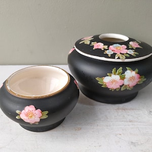 Puede incluir: Juego de cerámica negra de dos piezas con diseño floral. El juego incluye un cuenco pequeño y un recipiente con tapa. Ambas piezas presentan flores rosas y blancas con hojas verdes. Los interiores son de color crema.