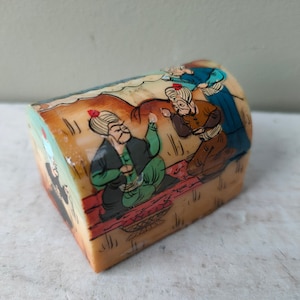Puede incluir: Una pequeña caja de madera con tapa curva, pintada con una escena de cuatro personas con ropa tradicional. La caja está decorada con detalles intrincados y colores vibrantes.