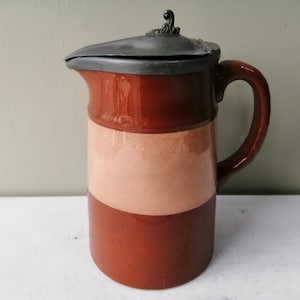 May include: A brown and cream ceramic jug with a pewter lid. The jug has a wide mouth and a handle. The lid has a decorative finial.