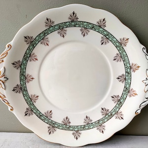 Op de afbeelding: Een witte keramische taartplateau met een gegolfde rand en een decoratieve groene en gouden rand. De plaat heeft twee handvatten.