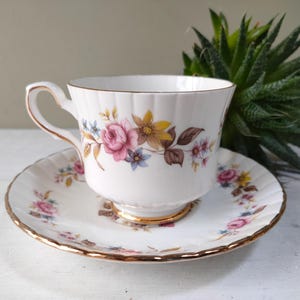 May include: A white teacup and saucer with a floral pattern in shades of pink, yellow, and brown. The cup and saucer have a gold trim.
