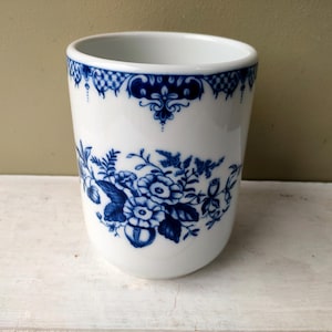 May include: A white ceramic tumbler featuring a deep blue floral pattern around the center and a decorative blue lace-like border along the top rim. The vessel has a smooth, cylindrical shape with a wide opening and a rounded base.