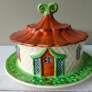 May include: A ceramic cottage-shaped butter dish with a green and white exterior, brown roof, and green base. The dish has a small, green, curly finial on top.