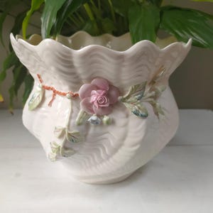 Könnte beinhalten: Ein weißer Keramiktopf mit einem gewellten Rand und einem Blumendesign. Der Topf zeigt eine rosa Rose mit grünen Blättern und einer Perlenkette.