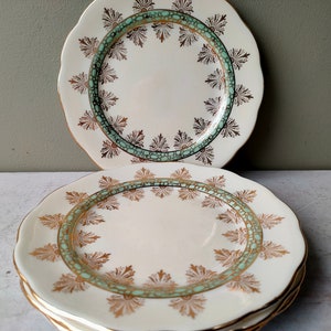 Op de afbeelding: Een set witte vintage borden met een geschulpte rand. Elk bord heeft een gouden rand en een decoratieve rand van gouden en teal bloemmotieven. De borden zijn op elkaar gestapeld, met twee borden zichtbaar in de afbeelding.
