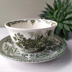 Puede incluir: Un juego de taza y platillo blancos con un diseño floral y de uvas verdes. La taza de té se encuentra sobre el platillo, con una planta verde en el fondo. El juego es de cerámica.