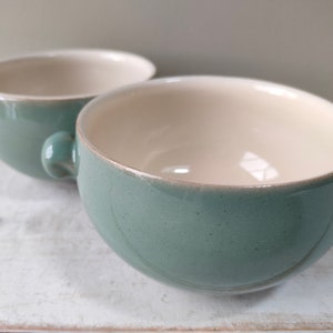 Op de afbeelding: Twee groene keramische soepkommen met een witte binnenkant. De kommen hebben aan elke kant twee handvatten.