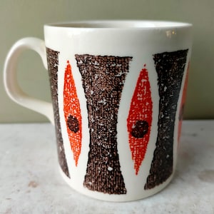 Könnte beinhalten: Eine weiße Keramiktasse mit einem geometrischen Muster in Orange-, Braun- und Schwarztönen. Das Muster zeigt eine Reihe von Diamantenformen mit einem strukturierten Hintergrund.