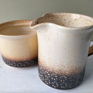 May include: A set of two ceramic cream and brown speckled sugar and creamer with a black lace design around the bottom.