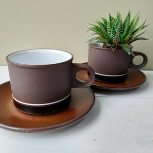 May include: Two matte brown ceramic cups with white interiors and matching saucers. Each cup features a thin white horizontal stripe near the base and a circular handle. One cup contains a small green succulent plant with pointed leaves.