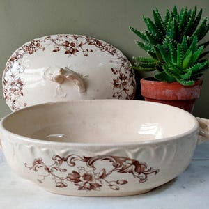 May include: Cream-colored antique serving bowl with a matching lid, adorned with brown floral patterns. The bowl has two handles and a decorative floral design around the rim. A small potted succulent plant sits nearby.
