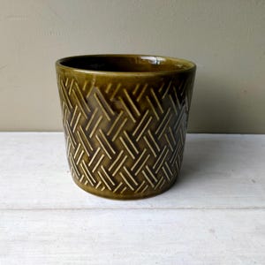 Op de afbeelding: Een cilindervormige keramische plantenpot met een diepe olijfgroene glazuurlaag. Het buitenoppervlak is voorzien van een herhalend reliëfpatroon van vlechtwerk, bestaande uit diagonale lijnen die een getextureerd, geweven uiterlijk creëren.
