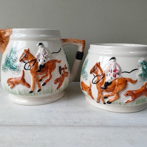 May include: Cream ceramic jug and sugar bowl set. The jug has a fox head spout and a handle with a black and brown design. Both pieces are decorated with a hunting scene of riders on horseback, dogs, and trees.