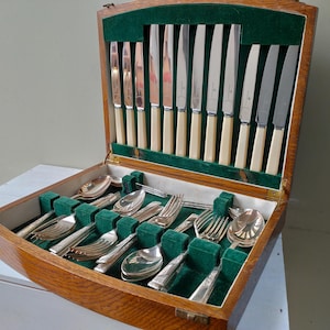 May include: An open wooden cutlery case with a green velvet lining, containing a full set of silverware. The set includes knives with cream-coloured handles, spoons, forks, and serving utensils. The case is open, displaying the contents.