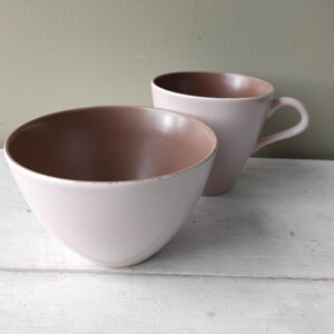 May include: A ceramic bowl and a matching mug with a smooth, light pink exterior and a contrasting warm brown interior. The bowl is rounded and open, while the mug features a curved handle on the side. Both pieces have a simple, clean design.