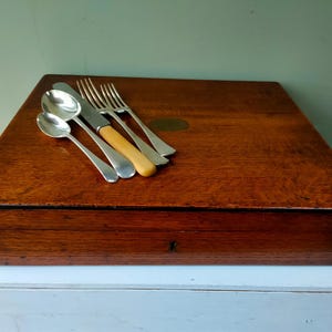 以下が含まれることがあります： 木製のカトラリーボックスの上に銀食器が置かれています。銀食器にはスプーン、フォーク、明るい色の柄のナイフが含まれています。ボックスは長方形で、濃い色の木材で作られています。