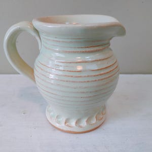 May include: A small, white ceramic pitcher with a light green glaze and a ribbed design. The pitcher has a rounded body and a flared lip.