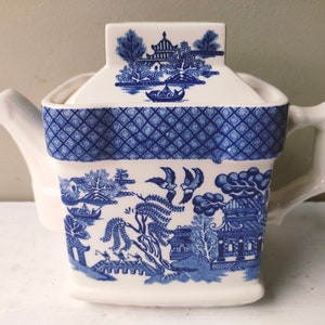 Puede incluir: Tetera de cerámica blanca con un diseño azul y blanco de patrón Willow. La tetera tiene un cuerpo rectangular, una tapa y un asa. El patrón azul representa un paisaje con edificios, árboles y pájaros. Una segunda tetera blanca es parcialmente visible.